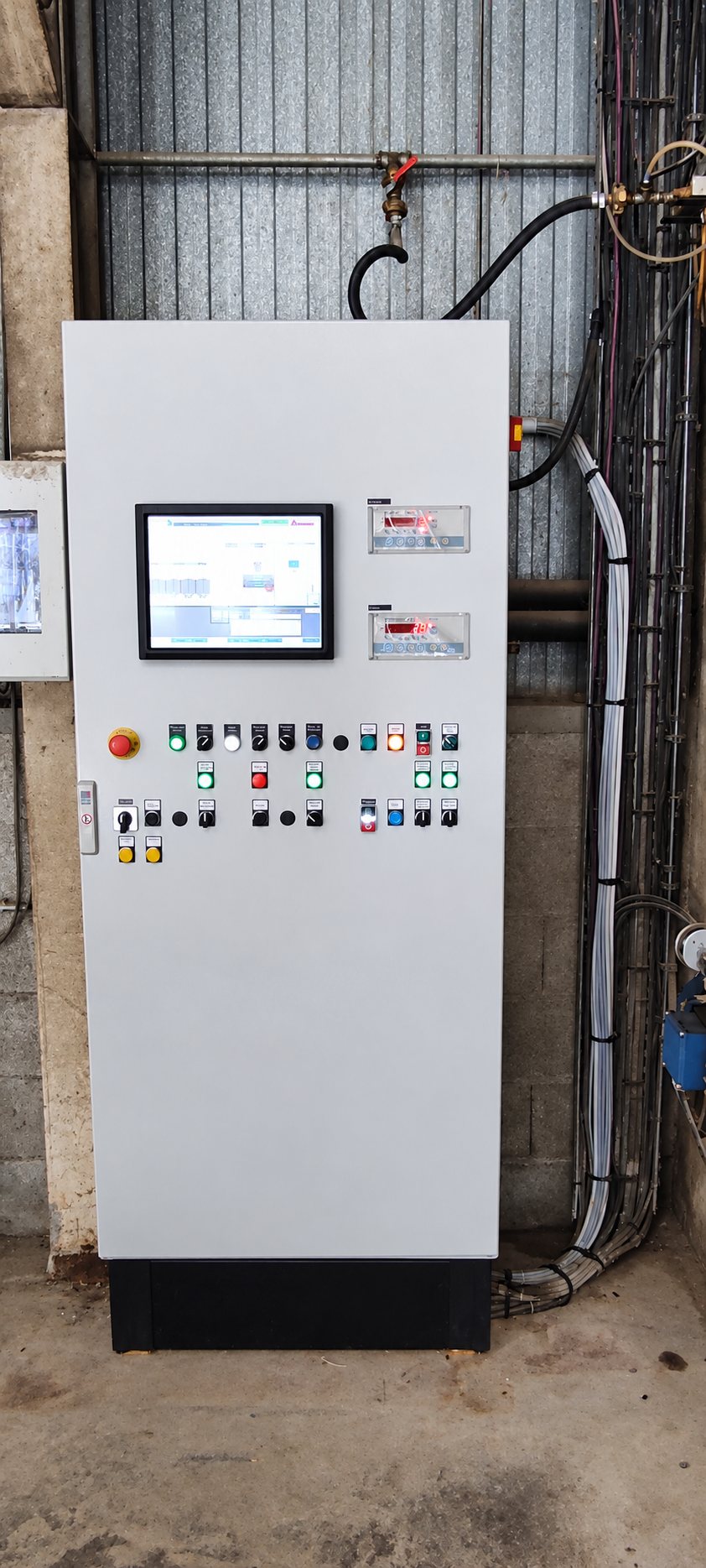 Armoire électrique grise équipée d’un écran tactile, de boutons lumineux et d’indicateurs, installée contre un mur dans un local industriel avec des câbles.