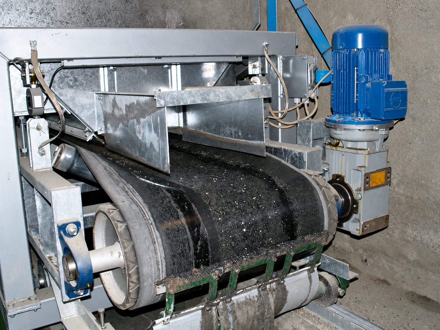 Convoyeur à bande industriel en acier galvanisé transportant une matière noire, entraîné par un moteur électrique bleu.