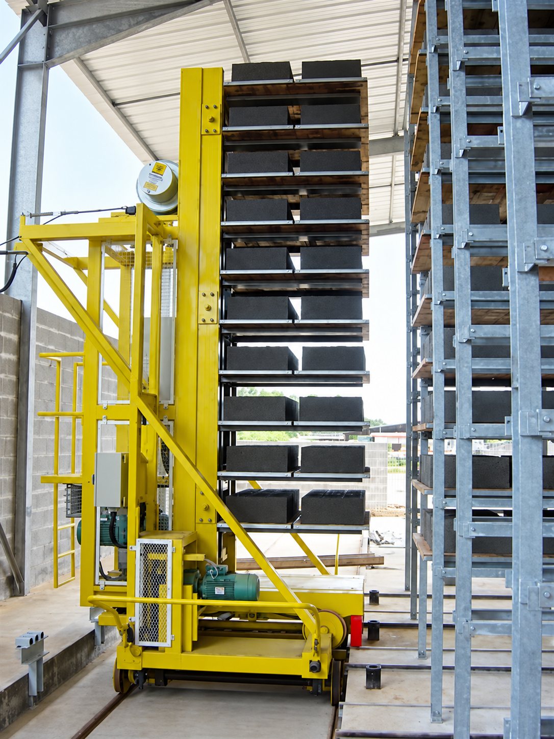 Grand élévateur industriel jaune chargé de rangées de blocs de béton gris, à côté d’un rack métallique galvanisé.